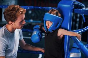 jovem treinador de boxe está ajudando o menino em roupas de proteção no ringue entre as rodadas foto