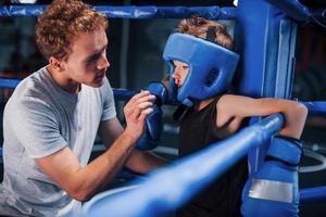 jovem treinador de boxe está ajudando o menino em roupas de proteção no ringue entre as rodadas foto