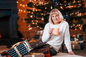 mulher alegre sênior sentada com um copo de bebida na sala com caixas de presente de natal foto