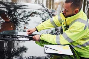 policial masculino de uniforme verde colocando lista de multas no veículo foto