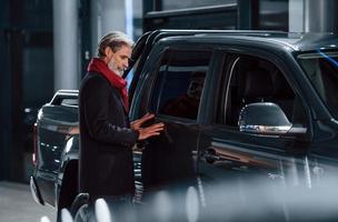 velho elegante com cabelos grisalhos e bigode procurando um carro novo dentro de casa no salão foto