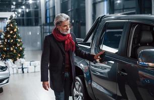 velho elegante com cabelos grisalhos e bigode procurando um carro novo dentro de casa no salão com árvore de natal foto