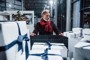 velho elegante com cabelos grisalhos e bigode embalando presentes em caixas de presente no carro. árvore de natal ao fundo foto