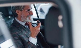 homem sênior elegante moderno com cabelos grisalhos e bigode falando ao telefone dentro do automóvel foto