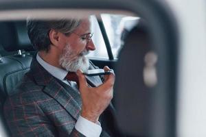 homem sênior elegante moderno com cabelos grisalhos e bigode falando ao telefone dentro do automóvel foto