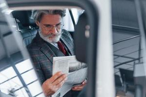 homem sênior elegante moderno com cabelos grisalhos e bigode lendo jornal dentro do carro foto