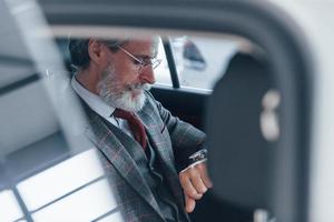 homem sênior elegante moderno com cabelos grisalhos e bigode olha para seus relógios dentro do automóvel foto