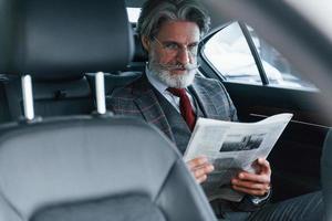 homem sênior elegante moderno com cabelos grisalhos e bigode lendo jornal dentro do carro foto