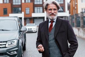 homem elegante sênior com cabelos grisalhos e barba em pé ao ar livre na rua perto de seu carro com as chaves na mão foto