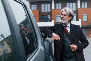 homem elegante sênior com cabelos grisalhos e barba, apoiando-se em seu carro e segurando as chaves foto