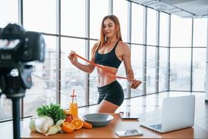 vlogger feminina com corpo esportivo em pé dentro de casa com fita métrica perto da mesa com comida saudável foto