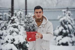 jovem bonito com casaco quente segurando pote vermelho com abeto ao ar livre durante o dia foto