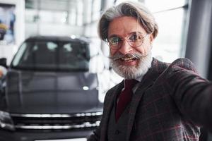 elegante homem sênior em roupas formais e com barba tira selfie contra carro novo moderno foto