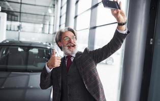 elegante homem sênior em roupas formais e com barba leva selfie contra carro novo moderno. mostrando o polegar para cima foto