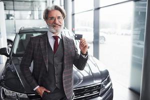 elegante homem sênior com cabelos grisalhos e barba fica contra o carro preto moderno com cartão de crédito na mão foto