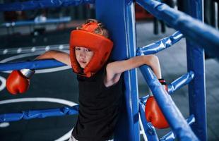 menino cansado em equipamento de proteção, apoiando-se nos nós do ringue de boxe foto