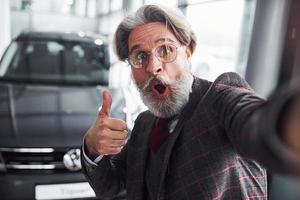 elegante homem sênior em roupas formais e com barba leva selfie contra carro novo moderno. mostrando o polegar para cima foto