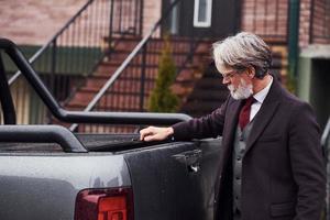 elegante homem sênior com cabelos grisalhos e barba está ao ar livre na rua perto de seu carro foto