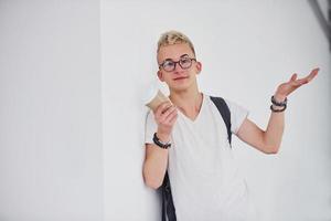 estudante em roupas casuais e com mochila fica dentro de casa contra a parede branca com um copo de bebida foto