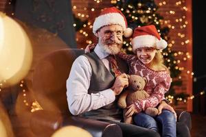 homem sênior alegre com cabelos grisalhos e barba sentado com a menina em chapéus de natal e ursinho de pelúcia foto