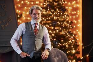 retrato de sênior elegante com cabelos grisalhos e barba na jaqueta e camisa branca está na sala de natal decorada foto