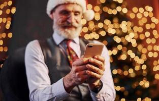 retrato de sênior elegante com cabelos grisalhos e barba sentado na sala decorada e com chapéu de natal foto