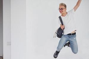 cara hipster feliz em roupas casuais e com mochila pulando dentro de casa contra a parede branca foto