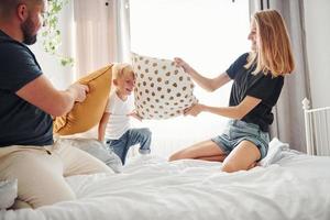 jovem casal com seu filho brincando de luta de travesseiros no quarto durante o dia foto