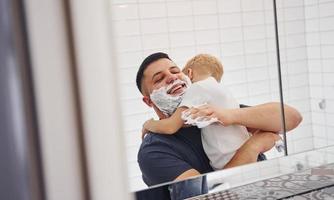 pai com seu filho está no banheiro se divertindo usando gel de barbear e olhando no espelho foto