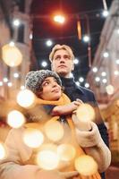 casal jovem positivo em roupas quentes, abraçando-se na rua decorada de natal foto
