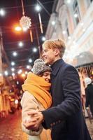 casal jovem positivo em roupas quentes, abraçando-se na rua decorada de natal foto