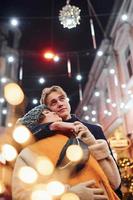 casal jovem positivo em roupas quentes, abraçando-se na rua decorada de natal foto