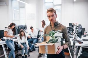 homem estiloso de óculos segurando caixa com planta na frente de seus colegas no escritório foto