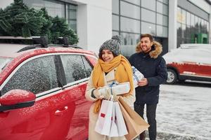 jovem casal com caixas de presente está perto de carro com árvore no topo. juntos ao ar livre no inverno foto