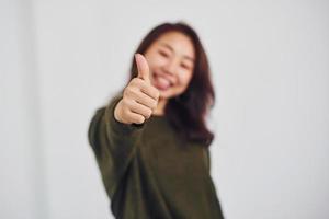 retrato de uma jovem asiática feliz que mostra o polegar para cima dentro de casa no estúdio contra um fundo branco foto