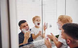 pai com seu filho está no banheiro se divertindo usando gel de barbear e olhando no espelho foto