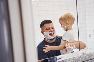 pai com seu filho está no banheiro se divertindo usando gel de barbear e olhando no espelho foto