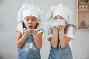 duas garotinhas de uniforme de chef azul sopram a farinha das mãos na cozinha foto