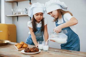 duas meninas em uniforme de chef azul derramando leite em copos na cozinha com biscoitos na mesa foto