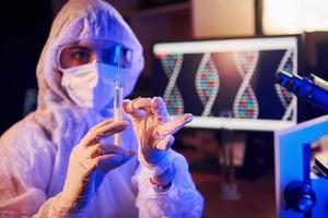 enfermeira com máscara e uniforme branco, segurando seringa e sentada em laboratório iluminado por neon com computador e equipamento médico em busca de vacina contra o coronavírus foto