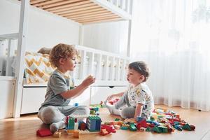 dois meninos se divertem dentro de casa no quarto com conjunto de construção de plástico. gato senta atrás deles foto