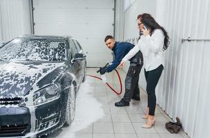 cliente do sexo feminino falando o que fazer. trabalhador masculino de uniforme lavando carro novo e moderno dentro de casa. concepção de serviço foto