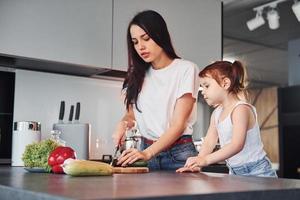 mãe com sua filha cortando legumes dentro de casa na cozinha foto