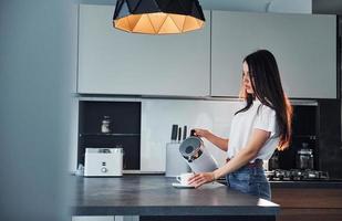 derramando leite fresco no copo. jovem morena linda em roupas casuais dentro de casa na cozinha durante o dia foto