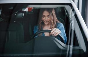 vista frontal da mulher positiva na camisa azul que fica dentro do novo carro novo. no salão de automóveis ou aeroporto foto