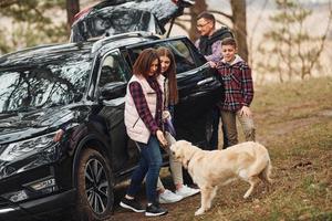 família feliz se diverte com seu cachorro perto de carro moderno ao ar livre na floresta foto