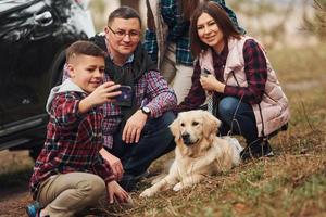 família feliz sentada e se divertindo com seu cachorro perto de carro moderno ao ar livre na floresta foto