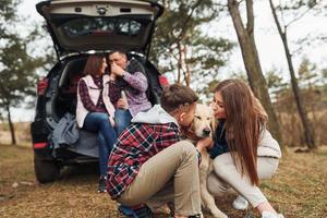 família feliz sentada e se divertindo com seu cachorro perto de carro moderno ao ar livre na floresta foto