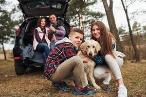 família feliz sentada e se divertindo com seu cachorro perto de carro moderno ao ar livre na floresta foto