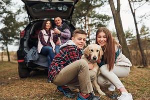 família feliz sentada e se divertindo com seu cachorro perto de carro moderno ao ar livre na floresta foto